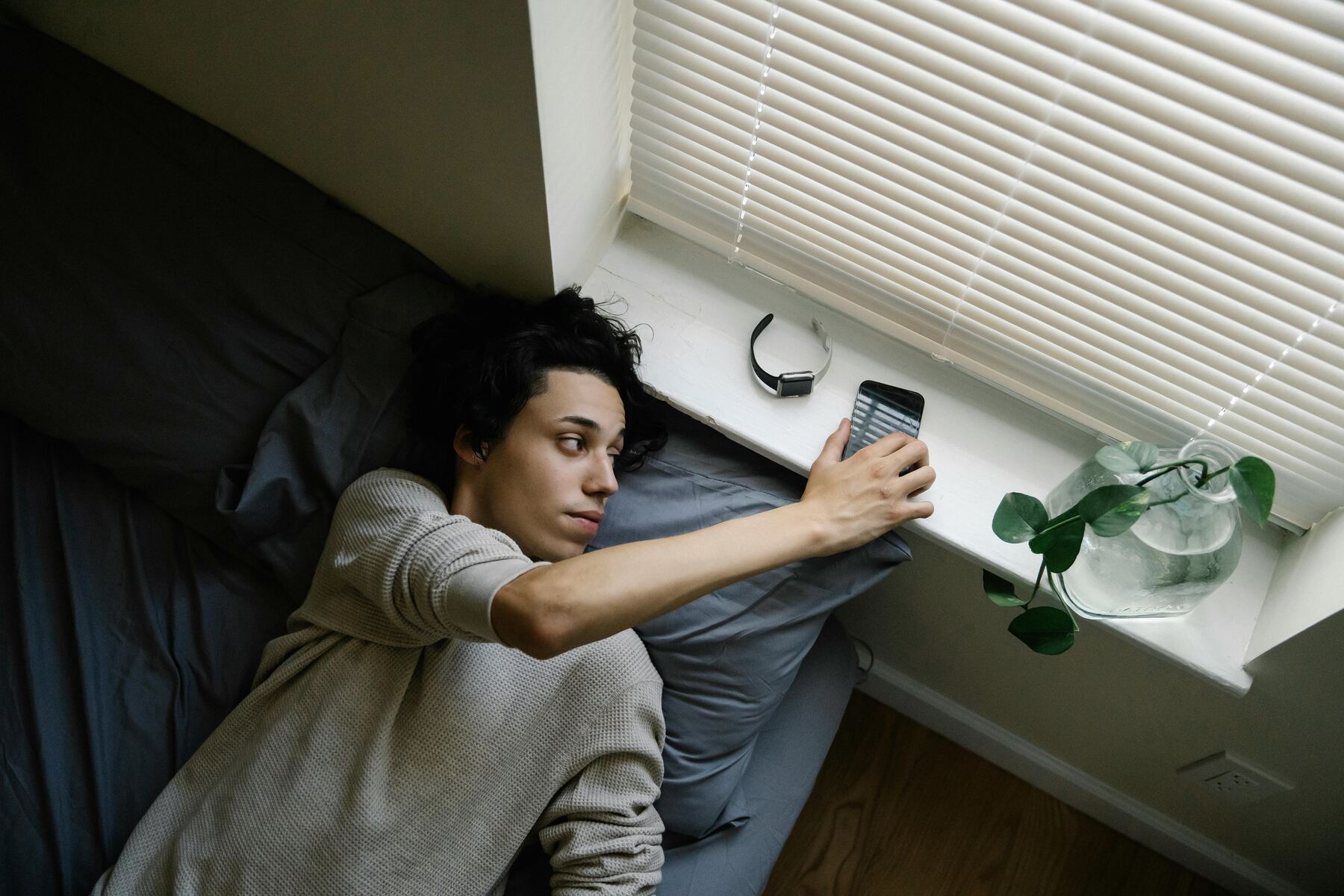 Young man lying in bed holding phone toward morning light — OnlyFans addiction symptoms and the phone-check loop